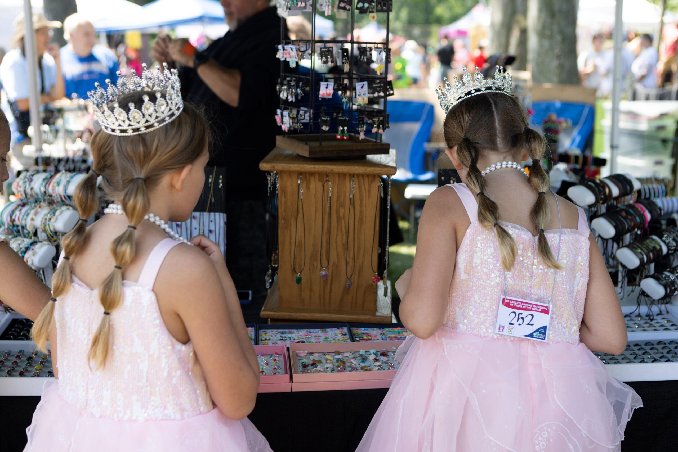 Twin girls dressed as princesses are seen from behind, admiring a vendor’s jewelry booth at the Twins Days festival.