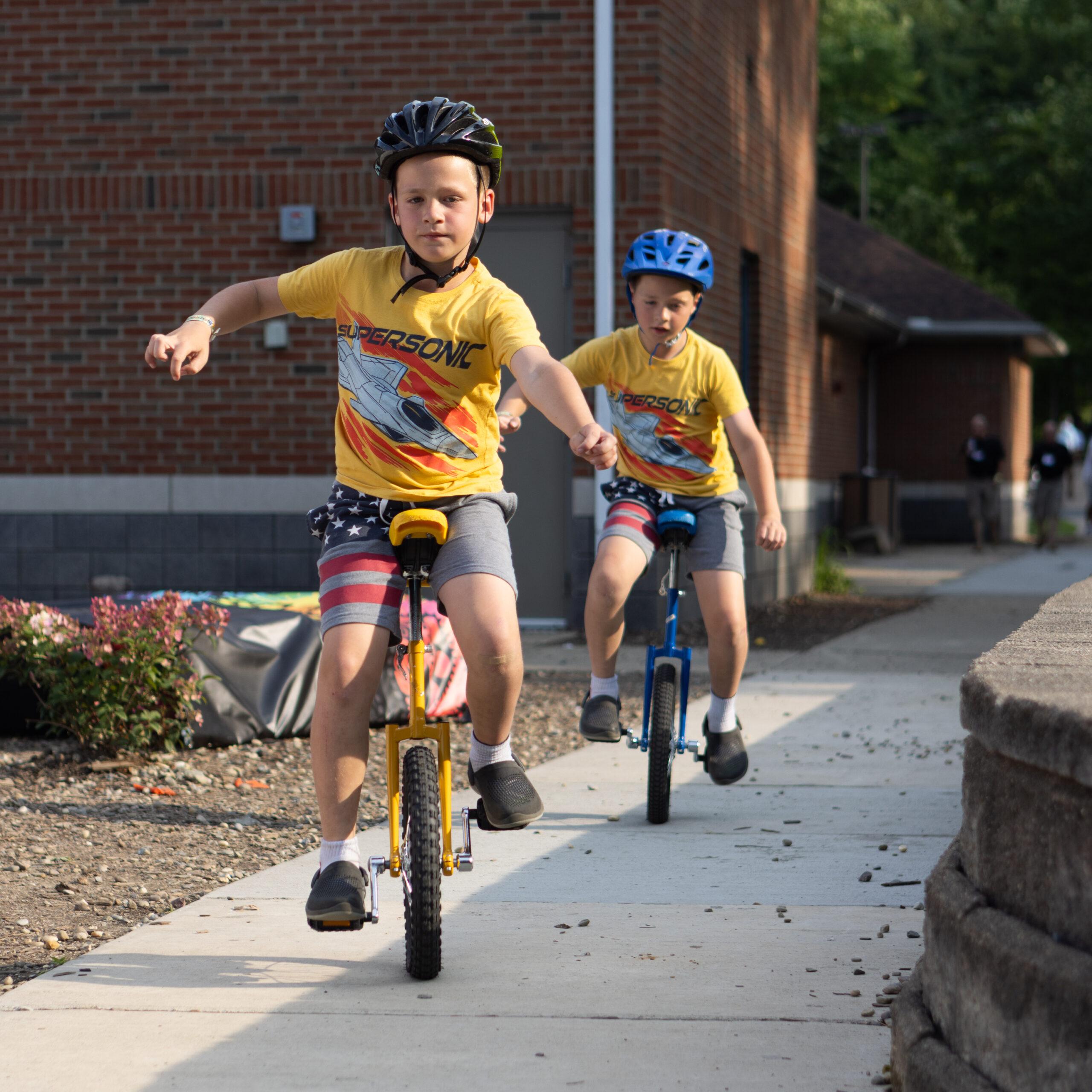 Twin boys on unicycles ride along a sidewalk at the Twins Days festival.