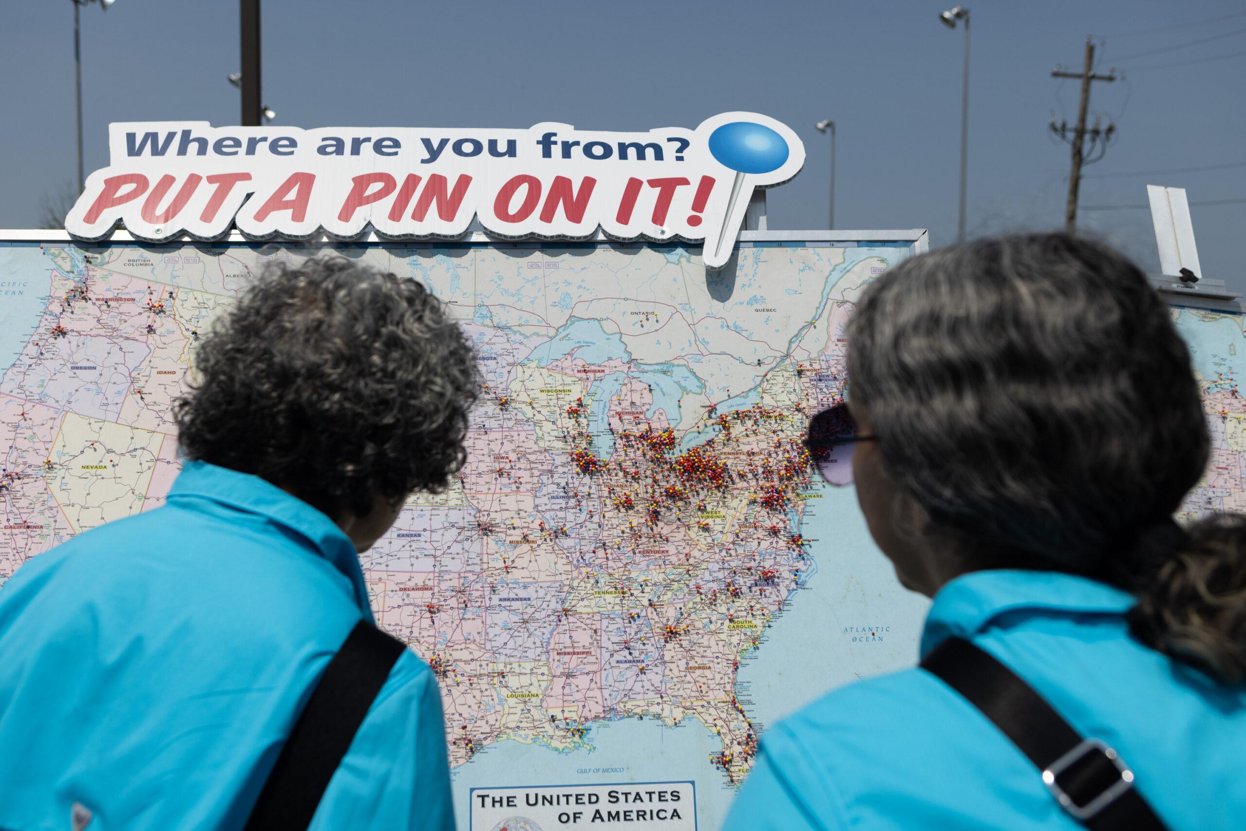 Twin women stand in front of a sign that reads “Where are you from? Put a pin on it!” above a map of the U.S. full of pins.
